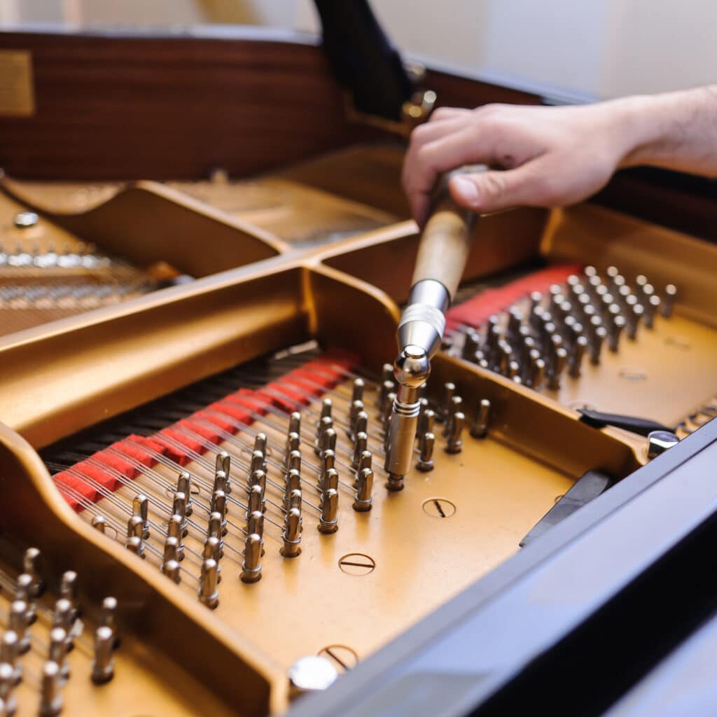 Main d'accordeur ajustant les chevilles d'un grand piano à l'aide d'un marteau. Le cadre doré, les cordes tendues et les feutres rouges sont visibles.