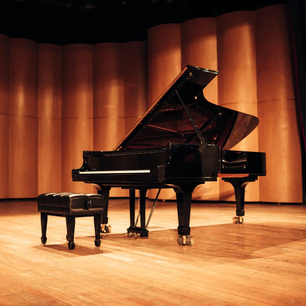 Grand Piano sur Scène : Ambiance Musicale Élégante de Concert Grand piano noir ouvert avec son banc en cuir sur scène en bois clair, devant des panneaux acoustiques bruns d'une salle de concert.