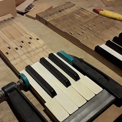 Clavier de piano en restauration, avec touches blanches et noires fixées par une pince. Le mécanisme en bois est visible, numéroté '50-54'.