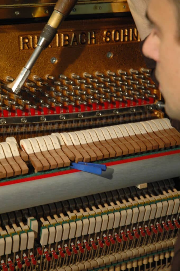 Réparation Piano Ibach : Ajustement des Marteaux Intérieurs Accordeur réglant un piano RÜLIBACH SOHN avec un outil, montrant les marteaux et la mécanique interne.